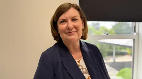 Wendy Reid has short straight brown hair. She is wearing a dark blue suit jacket over a patterned top. She is standing in an office in front of a window.
