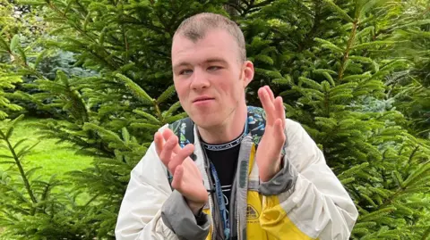 Billy is a young man with dark brown hair. He is wearing a cream jacket with a bit of yellow and is clapping. Behind him is a very green tree.