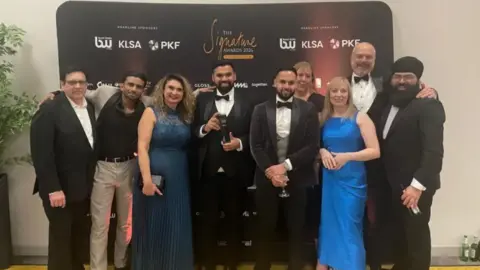 Nine men and women wearing various forms of formal evening dress pose for a picture at an awards ceremony. The man in the middle, in a dinner jacket and bow tie, is holding a black trophy. The group is standing in front of a black branded media wall featuring several sponsor names alongside the event name, The Signature Awards 2026. 