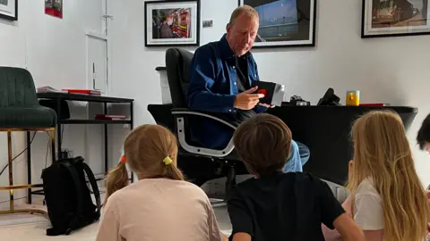 The image shows a man sat on a chair talking to a group of five children who are all sat on the floor. Framed pictures can be seen on the wall in the background.