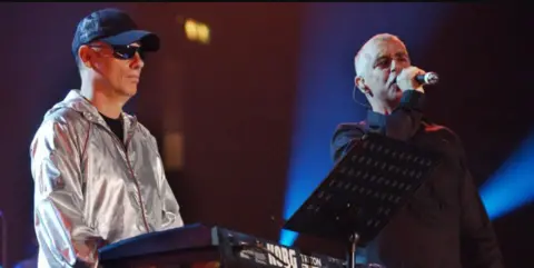 PA Media The Pet Shop Boys on stage in 2022, Chris Lowe, on the left is wearing a silver jacket, cap and glasses as he stands next to a keyboard. Neil Tennant stands close to him holding a microphone to his mouth.