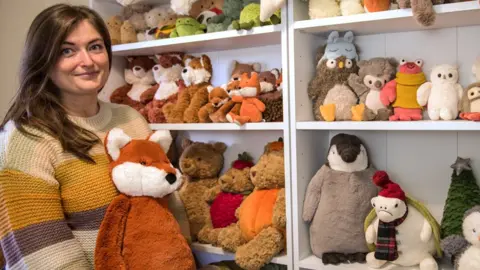 Amanda Hope stands holding a toy fox next to her collection of Jellycat toys stacked on shelves