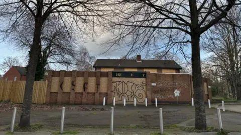 A disused pub with boarded windows and graffiti on its walls. There are two leafless winter trees in front of it. The building is abandoned, with metal bollards and leaves in front of it. 
