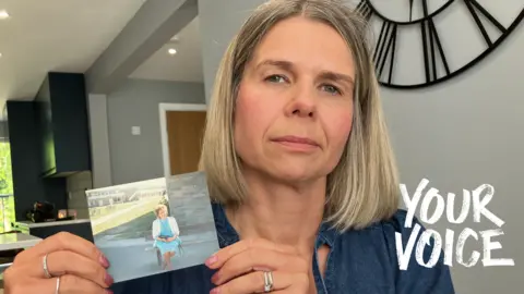 Michelle holds a small printed photograph with both hands. She is wearing a loose, long-sleeved top in a muted blue tone. The sleeves have gathered cuffs, and the fabric appears soft and casual. On her fingers, several rings are visible, including a band on one finger and another ring with a darker stone on a different finger. She is holding a small rectangular photograph at chest level. The photograph shows her mother sitting on a bench in sunlight, wearing a light blue outfit, with a building and grassy area in the background.