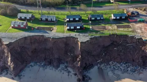 A bird's eye view of dark red holiday chalets dotted along a coastline. The coast is eroding with bits of rock seen to the right of the photo on the sand below, with other items fallen half way down the cliff.