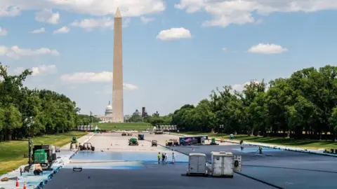Workers and work trucks in the drained Lincoln Memorial reflecting pool, which is partially painted blue, with the Washington Monument in the background