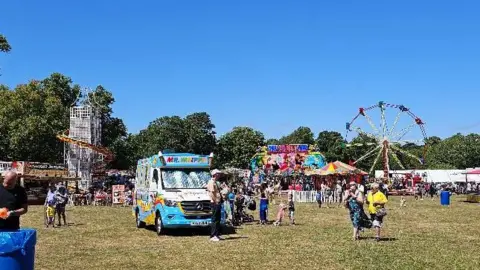 Graham's Vista A crowded grassy area with an ice cream van in the foreground and a helter skelter and big wheel in the distance