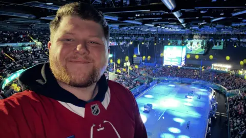 John McAllister in a stadium at the Winter Olympics. Behind him is an ice hockey rink 