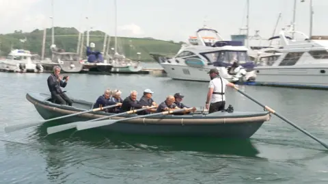 Six rowers row a grey boat in a harbour