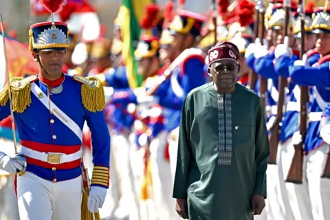 Evasristo Sa/AFP/Getty Images Nigeria's President Bola Tinubu, wearing a green outfit and maroon hat, reviews ceremonial guards dressed in white trousers, blue jackets and elaborate helmets  at Planalto Palace, Brasília, Brazil - Monday 25 August 2025. 