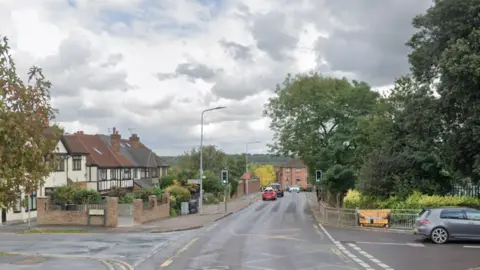 A street view image of a yellow box junction on Palmerston Road. There is a crossing with traffic lights up ahead and houses on the left.
