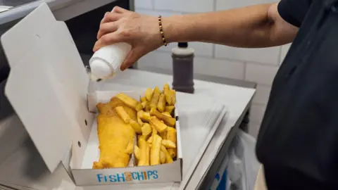Over the shoulder shot of a worker holding a salt pot over a portion of fish and chips. The food is inside a carboard box with a lid.