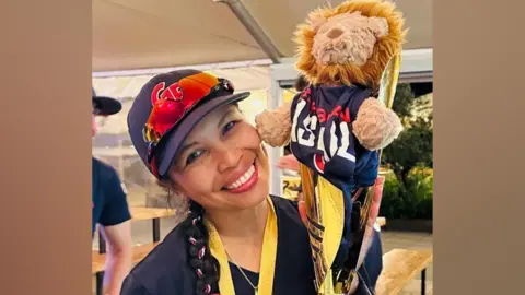 A woman with a blue baseball cap and dark blue tshirt stands smiling and holding gold cup trophy with a soft toy lion inside. Her dark brown hair is tied into a plait with red, white and blue ribbons and hangs over her shoulder.