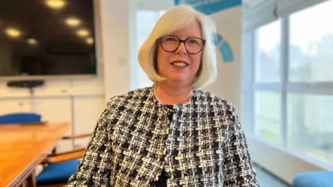 BBC A women with white hair that comes down to her jaw. She is wearing round glasses and smiling. The women is wearing a black and white patterned jacket and a single strand of pearls around her neck.