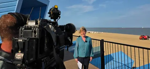 A cameraman in the foreground filming Look East presenter Susie Fowler-Watt, who is looking direct at the camera in front of an empty beach with blue skies in the background