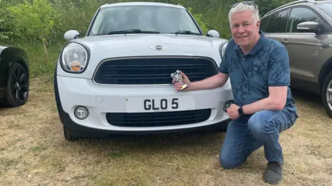 Neal Bircher Neal Bircher poses on bent knee in front of his white Mini that has a 'GLO 5' number plate