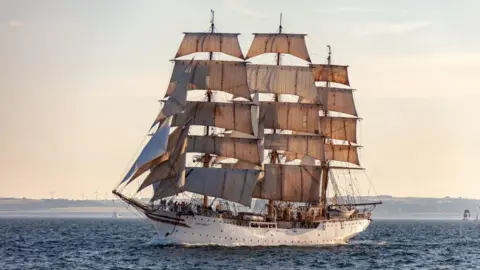 Tall cream-coloured ship - Sorlandet - with masts and beige sails at sea, with land and wind turbines in the distance.