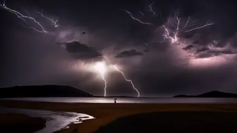 FLLOYD MALLON A man with an umbrella stands at the edge of a beach in Fingal Bay, New South Wales during a lightning storm