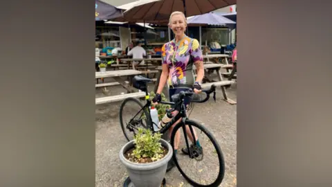 Julie Rose Julie is standing outside a cafe holding her bicycle against a large plant pot. There are lots of picnic tables and parasols behind her on the tarmacked ground. She is wearing a pastel-coloured, patterned cycle shirt and is smiling to camera