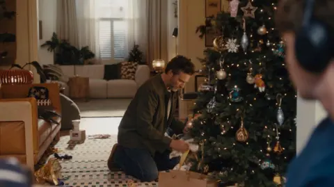 A father wearing a khaki shirt, a white t-shirt and blue jeans opening a present under a Christmas tree decorated with classic baubles. His son is watching, wearing a blue jumper and black headphones. 

