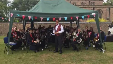 Enderby Band Image shows President of the organisation, Garry Sleath, standing in front of a band. He is wearing a red waistcoat over a white shirt, black trousers and black shoes. The other band members are sat behind him in a large semi-circle, all dressed in black suits with a red opening on the jackets. They're outside on some grass and underneath a dark green marquee with ribbons round the top edges. 