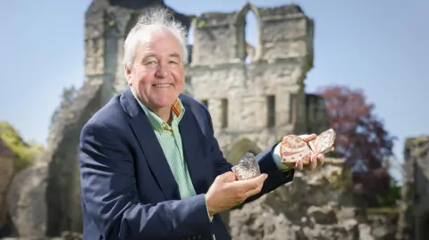 A man with white hair a blue jacket and pale green shirt holding small tiles in both hands with a ruined stone building behind him