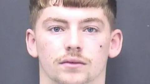 A mugshot of Betteridge. He has blue eyes, a light brown fringe, and thin moustache.