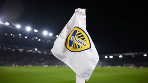 A corner flag with the Leeds United badge on, set against the backdrop of a floodlit football stadium.