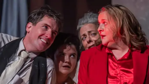 Still photography from Faust-ish play - a woman with blonde hair, in red silk pyjamas, leaning against a table. On the table a man with a grey suit is lying across it smiling. Beside her two women, both in grey bedwear, stand close to her, looking shocked. The staging is minimal, with several sheets draped across a red brick background.