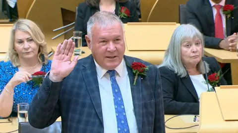 Davy Russell in the Holyrood chamber, wearing a suit with a right hand raised.