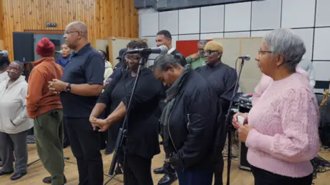 Homens e mulheres ficam ao lado dos microfones no estúdio. Eles estão vestindo blusas e calças e alguns usam casacos e óculos.