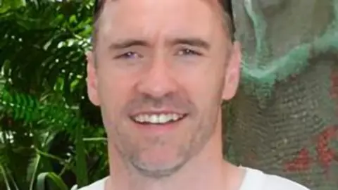 PA Media Paul Doyle has short dark greying hair with sunglasses on his head and is wearing a white T-shirt