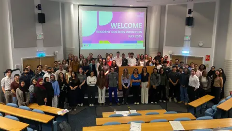 SaTH More than 100 people are standing in a semi circle in front of an induction sign in a lecture theatre. 