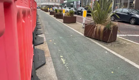 Red barriers beside a cycle lane in Belfast city centre. Large potted plants and parked cars are also visible in the picture