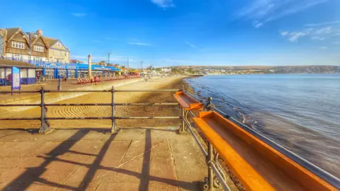 Susan Ward On the left is the beach with amusement shops all along the promenade and on the right is the sea, which is still, with just a ripple on the water. The sky is clear blue and there are railings on the path at the front of the photograph on this sunny day.