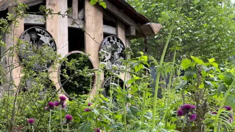 A wooden structure with large round windows and a pitch roof behind flowers and green foliage
