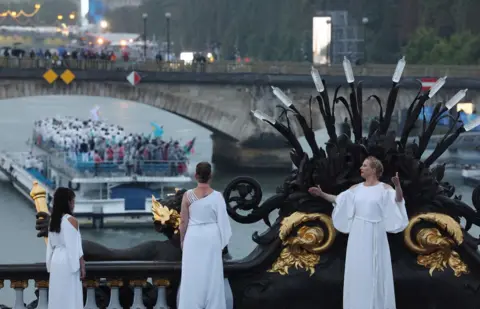   Pilar Olivares/Reuters Performers dressed in white robes stand on Pont Alexandre III during the opening ceremony of the Paris 2024 Olympics, as a boat passes beneath