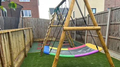 Part of a garden fenced off. There is artificial grass, specialist flooring and an adapted wooden play set with a large multi-coloured swing.