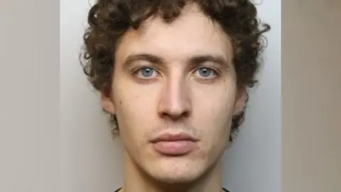 A mugshot of a man with short, curly brown hair. He is staring blankly at the camera in front of a grey background.