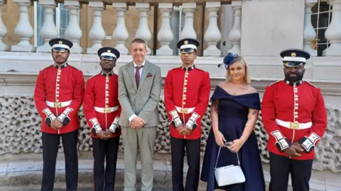 Dave Poulton Dave wearing a beige suit, white shirt and a red tie and pocket square, standing with four soldiers in red coats, and his daughter who is in a blue dress wearing a blue fascinator hat.