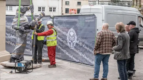 Two men working on a statue which is wrapped in dark-coloured cling film and has green straps attached to it. Four men to the right watch on. Behind them are barriers with Motorhead branding.