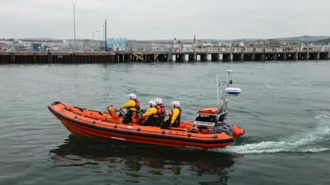 Weymouth inshore lifeboat with four crew members on board wearing yellow waterproofs, red lifejackets and white helmets. The boat is an orange rib and is sailing into Weymouth Harbour with Weymouth Pier in the background and the seafront behind it in the distance.