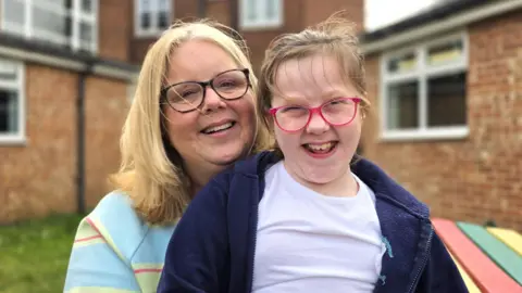 Claire and Katie, sitting outside and smiling at the camera. On the left Claire is wearing black rimmed glasses and and a light blue and green jumper. Sitting on her lap is Katie, who has pink rimmed glasses, whilst wearing a dark blue fleece over a light violet t-shirt.