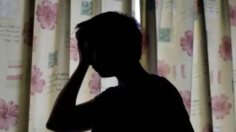 A silhouette of a woman holding her hand against her head. She is standing in front of floral curtains which are drawn. 