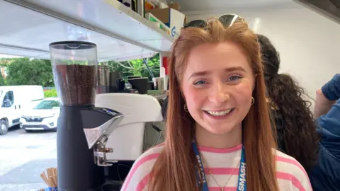 A young woman with red hair smiling at the camera from inside a mobile food van with a coffee machine and other people in the background.