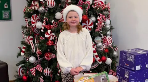 Family A young girl surrounded by toys and other presents such as chocolate. She is smiling at the camera. She is wearing a white jumper, leopard legging and a Christmas hat. There is a Christmas tree with lots of decorations behind her.
