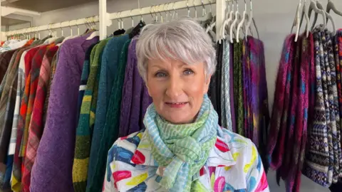Sandra, who is inside her shop, has short grey hair and is wearing a colourful green and blue patterned scarf and a brightly coloured top. She is smiling as she stands in front of a rack of brightly coloured clothes on hangers.