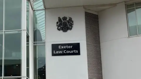 BBC Exterior of Exeter Crown Court. The main focus of this cropped image is the Exeter Law Courts sign on the front entrance of the building. To the left of the sign are large glass windows.