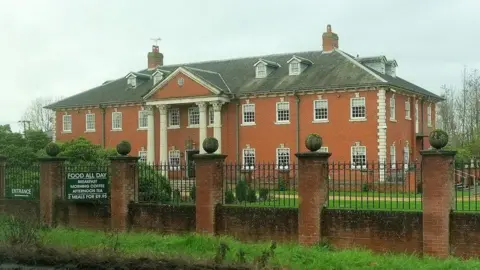 Elme Hall, pictured from the A47. It's a large hotel with four columns at the front. In the foreground is a wall made up of bricks and metal railings.