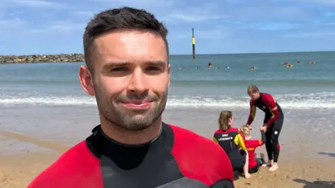 Qays Najm/BBC Ted Morgan a man standing on the sand of the beach at Sea Palling. He has short brown hair and is looking directly at the camera and smiling. Behind him is the beach, the sea and a blue sky.
